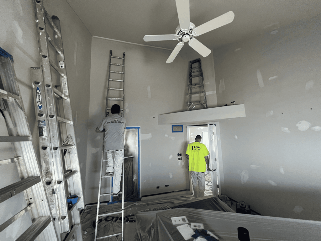 Worker sanding drywall from a ladder in a tall room prepared with floor protection