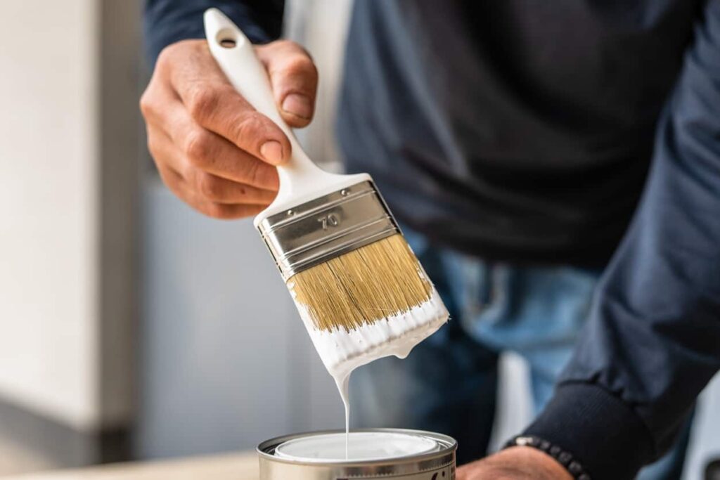 Hand holding a paintbrush above an open can of white paint